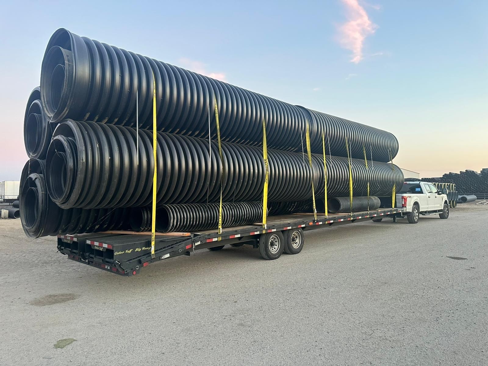 White truck transporting large black pipes on a flatbed trailer in an industrial area.